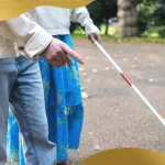 Visually impaired person using a white cane while walking with assistance on a park pathway, representing blindness, mobility support, and vision impairment awareness.