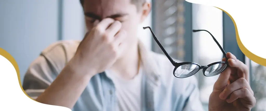 Man experiencing eye strain and blurry vision while holding eyeglasses, a common symptom of uncorrected refractive errors or astigmatism