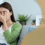 Woman sitting on a couch rubbing her eyes in discomfort while using a laptop, indicating digital eye strain or vision problems