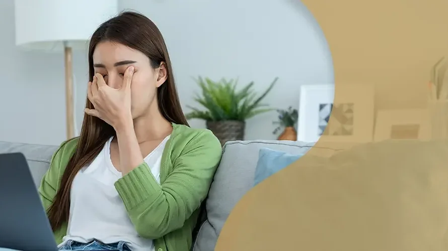 Woman sitting on a couch rubbing her eyes in discomfort while using a laptop, indicating digital eye strain or vision problems