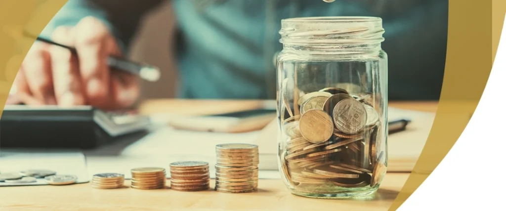 Stacked coins and a jar of savings on a desk with a person calculating expenses in the background, symbolizing budgeting and cost planning for LASIK surgery